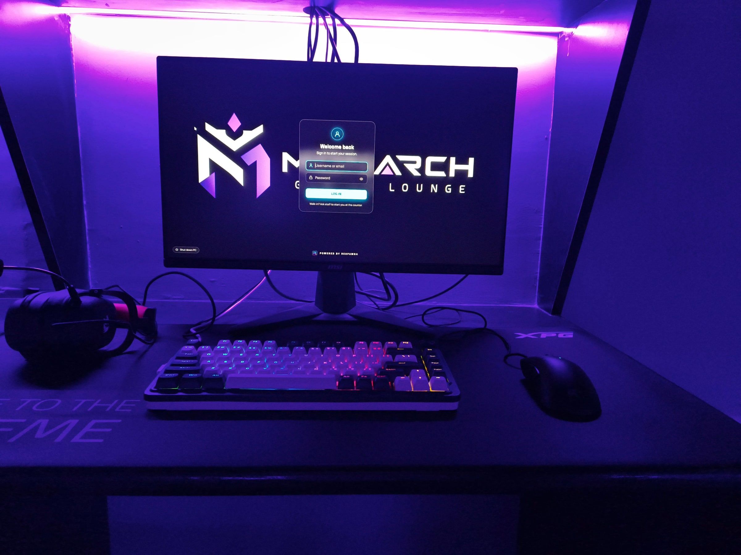 A PC station at Monarch: the monitor shows the Monarch sign-in screen, with a backlit keyboard, mouse and headset under purple light.